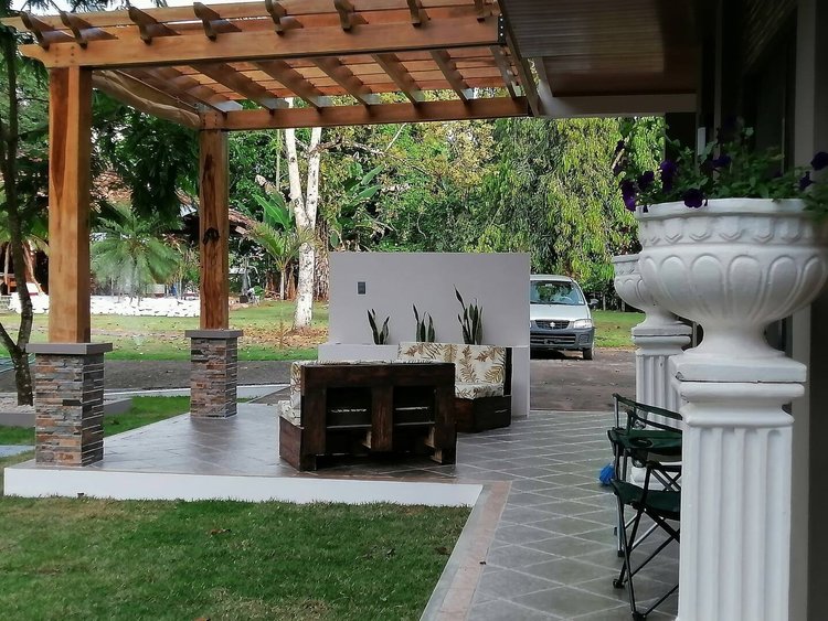 A patio area with a wooden pergola, brick pillars, and outdoor seating including a bench with floral cushions. There are large potted plants and trees in the background, with a car parked nearby.