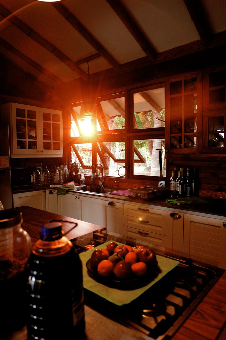 Kitchen with sunlight streaming through windows, showing a bowl of fruit on a stovetop, cabinets, and bottles on the counter.