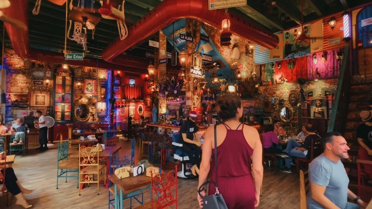 Interior of a lively restaurant decorated with colorful, eclectic artwork and ornaments, featuring patrons seated at tables and a woman with a ponytail in a red dress walking through.