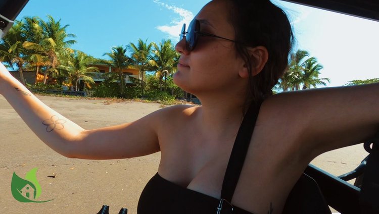 Woman with sunglasses sitting in a golf cart on a beach, with palm trees and a house in the background, on a sunny day.