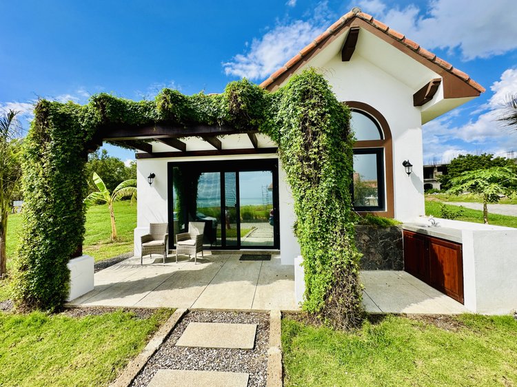A modern house with white walls and a red tile roof, featuring a front porch with two chairs and lush green vines growing over a pergola, under a bright blue sky.