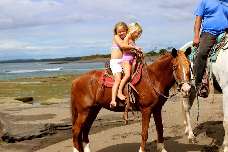 Two young girls riding a brown horse on a beach, smiling and hugging, with a person on a white horse nearby. The sky is partly cloudy, and the beach has rocks and sand.