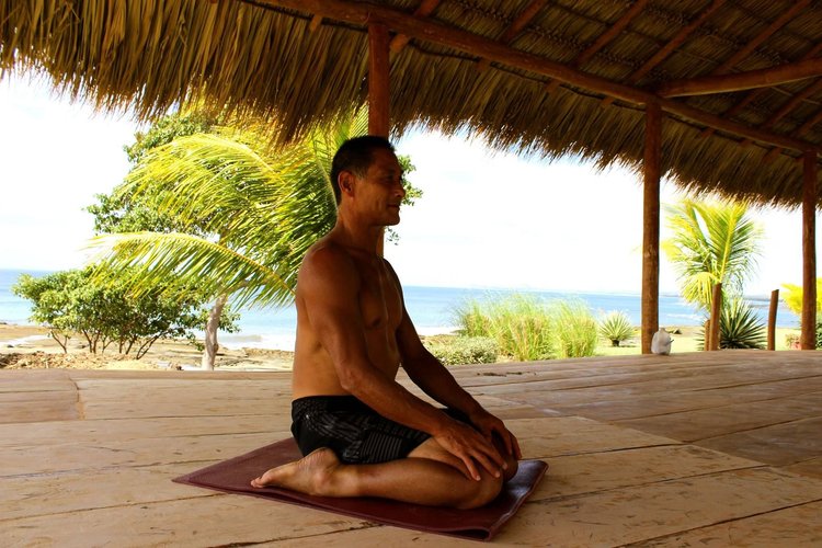 A man practicing yoga on a mat under a thatched roof with a beach view, palm trees, and ocean in the background.