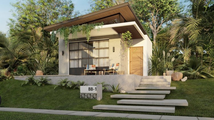 Modern small house with a sloped roof, large front window, and wooden door, surrounded by lush green plants and trees, with a patio featuring two chairs and a table.